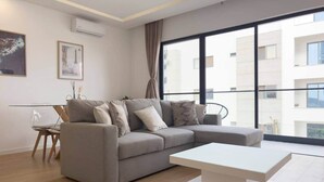 Uma sala de estar moderna com um sofá cinza, uma mesa de centro branca e uma janela grande com vista para edifícios.