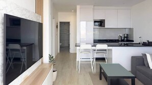 Una cocina moderna con armarios blancos, una encimera negra y un microondas de acero inoxidable. Hay una zona de comedor con sillas blancas y una mesa pequeña.
