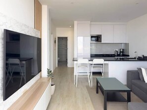 A modern kitchen with white cabinets, a black countertop, and a stainless steel microwave. There is a dining area with white chairs and a small table. - Cozy 2 bdr aprt, rooftop pool - LCGR (Praia)