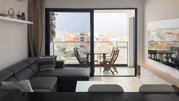 Una sala de estar moderna con un sofá, una mesa de comedor con sillas y un amplio ventanal corredera que da a un balcón exterior.