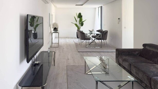 A modern living room with a flat-screen TV, a glass coffee table, a dark sofa, and a dining area with chairs and a plant. - 2-bdr bright apt cidadela central street - LCGR (Praia)