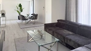 A modern living room with a glass coffee table, a grey sofa, a dining area with chairs, and a plant. - 2-bdr bright apt cidadela central street - LCGR (Praia)
