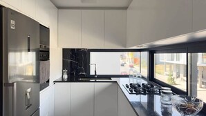 A modern kitchen with a stainless steel refrigerator, white cabinetry, a black marble backsplash, and a view of the city through large windows. - 2-bdr bright apt cidadela central street - LCGR (Praia)