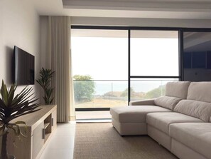 A modern living room with a sectional sofa, a flat-screen TV, a wooden console, and a large window with a view. - Jorge: Palmarejo Baixo (Praia)
