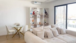 Ein modernes Wohnzimmer mit einer Couch, einem Glastisch, Stühlen, einem weißen Bücherregal und einem großen Fenster.