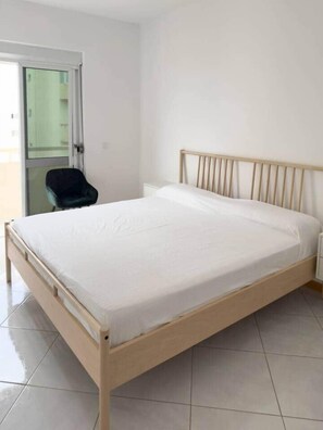 Een slaapkamer met een houten bed, witte muren, een stoel en een deur naar een balkon.