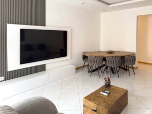 Een moderne woonkamer met een grote flatscreen televisie aan de muur, een ronde eettafel met grijs beklede stoelen en een houten salontafel met decoratieve objecten.