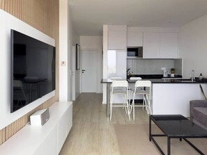 Une cuisine moderne avec des armoires blanches, un comptoir noir et un micro-ondes encastré. On y trouve un coin repas avec une table et des chaises blanches, ainsi qu’un téléviseur à écran plat fixé au mur.