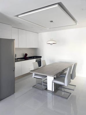 Une cuisine moderne avec des meubles blancs, un réfrigérateur en acier inoxydable, une cuisinière noire et une table à manger avec des chaises blanches.