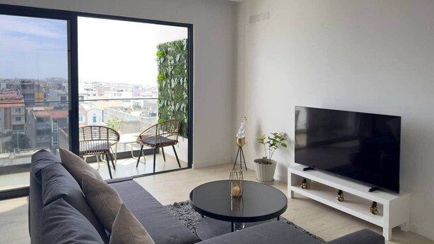 A modern living room with a large window, a sofa, a round coffee table, a TV on a white stand, and a small balcony with a chair. - Spacious 2 bdr aprt, rooftop pool - LCGR (Praia)