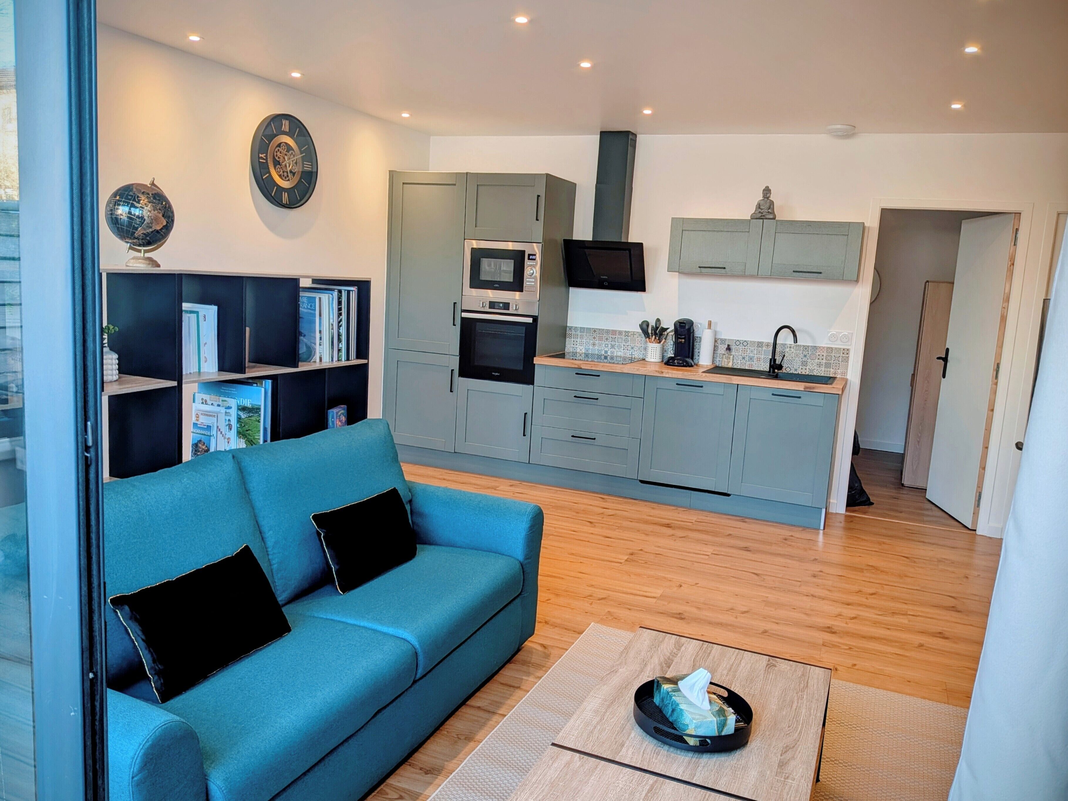 Una sala moderna con un sofá azul, una mesita de centro y una cocina con armarios grises, un microondas y un televisor.