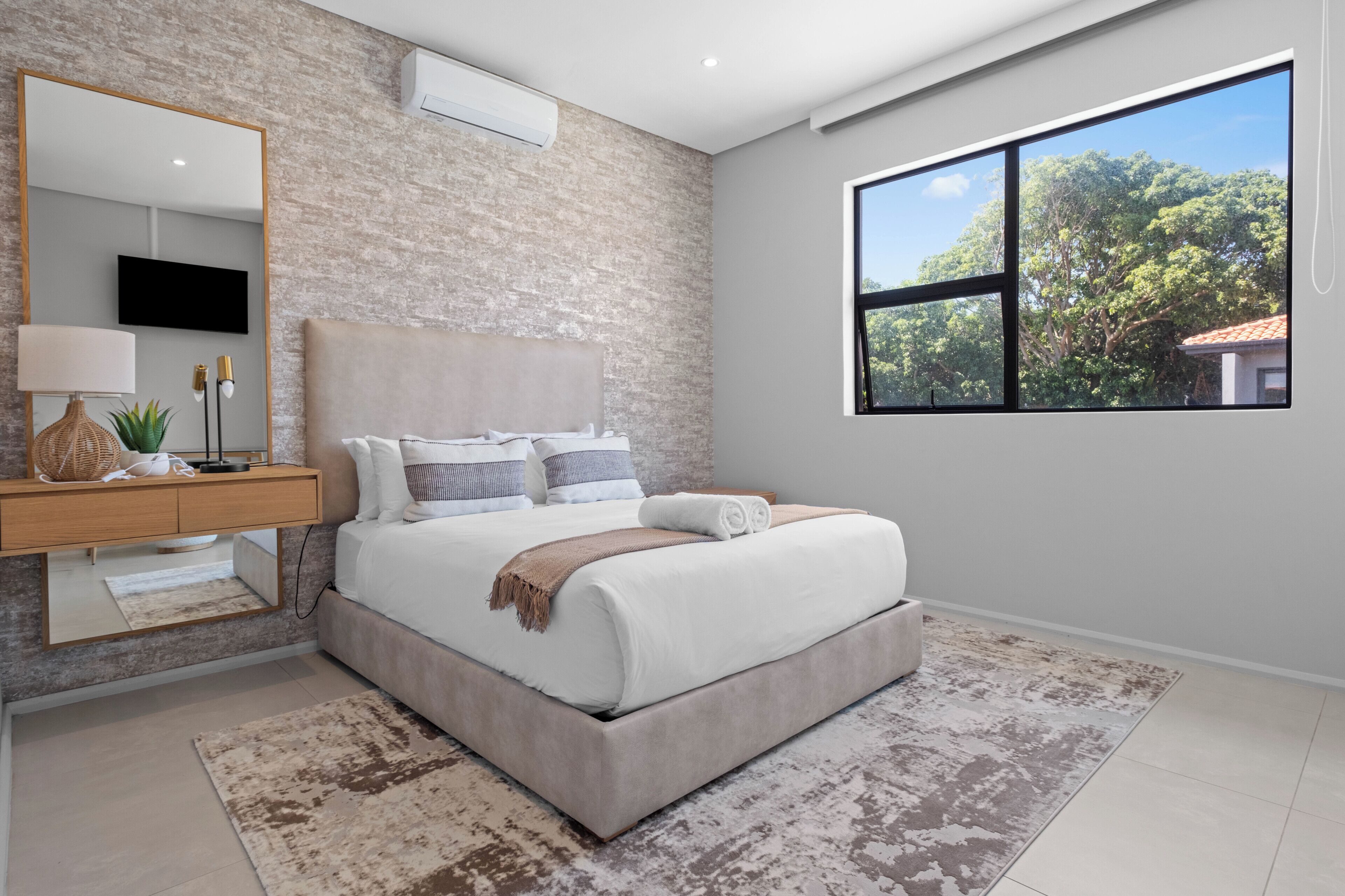 A modern bedroom with a large bed, a bedside table, a mirror, and a large window with a view of trees.