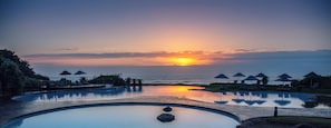 A swimming pool with lounge chairs and umbrellas, overlooking the ocean at sunset.