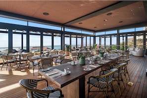 A spacious outdoor dining area with multiple tables and chairs, large windows offering a view of the city, and a wooden deck.