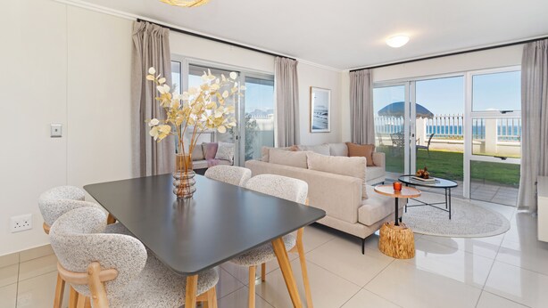 A modern living room with a dining table, sofa, and large windows offering a view of the outdoors. - Beachfront Apartment | Table Mountain Views (Cape Town)
