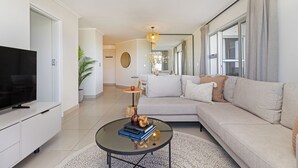 A modern living room with a sofa, a coffee table with books and a candle, a TV on a white cabinet, and a large window with curtains. - Beachfront Apartment | Table Mountain Views (Cape Town)