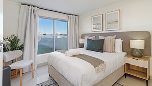 A modern bedroom with a large bed, a nightstand, a chair, a window with curtains, and two framed pictures on the wall. - Beachfront Apartment | Table Mountain Views (Cape Town)