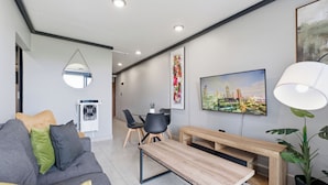 A modern living room with a sofa, a wooden coffee table, a TV, a lamp, and a dining area with chairs and a table. - Stylish 2BR @ The Onyx | Rooftop Pool and Braai (Umhlanga)