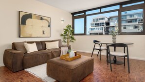 Een moderne woonkamer met een bruine bank, een bijzettafeltje met dienblad, een eethoek met een kleine tafel en stoelen, en een groot raam met uitzicht op een meer verdiepingen tellend gebouw.