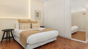 Een slaapkamer met een bed, een klein krukje, een spiegel en een kledingkast.