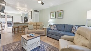 Ein Wohnzimmer mit einer Couch, einem Couchtisch, einem Fernseher, einem Deckenventilator und einer Küche im Hintergrund.