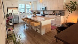 A modern kitchen with white cabinets, a stainless steel refrigerator, a large island with bar stools, pendant lighting, and a potted plant.