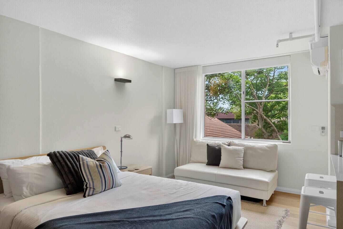 Un dormitorio con cama, sofá, ventana con cortinas, mesita de noche y lámpara.