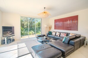 Ein modernes Wohnzimmer mit großem Flachbildfernseher, einer Couch, einem Couchtisch und einem großen Fenster mit Blick auf Grünflächen.