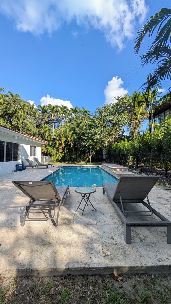 A swimming pool with lounge chairs and a table, surrounded by palm trees and a house. - Coral Gables Vacation House with Pool (Coral Gables)