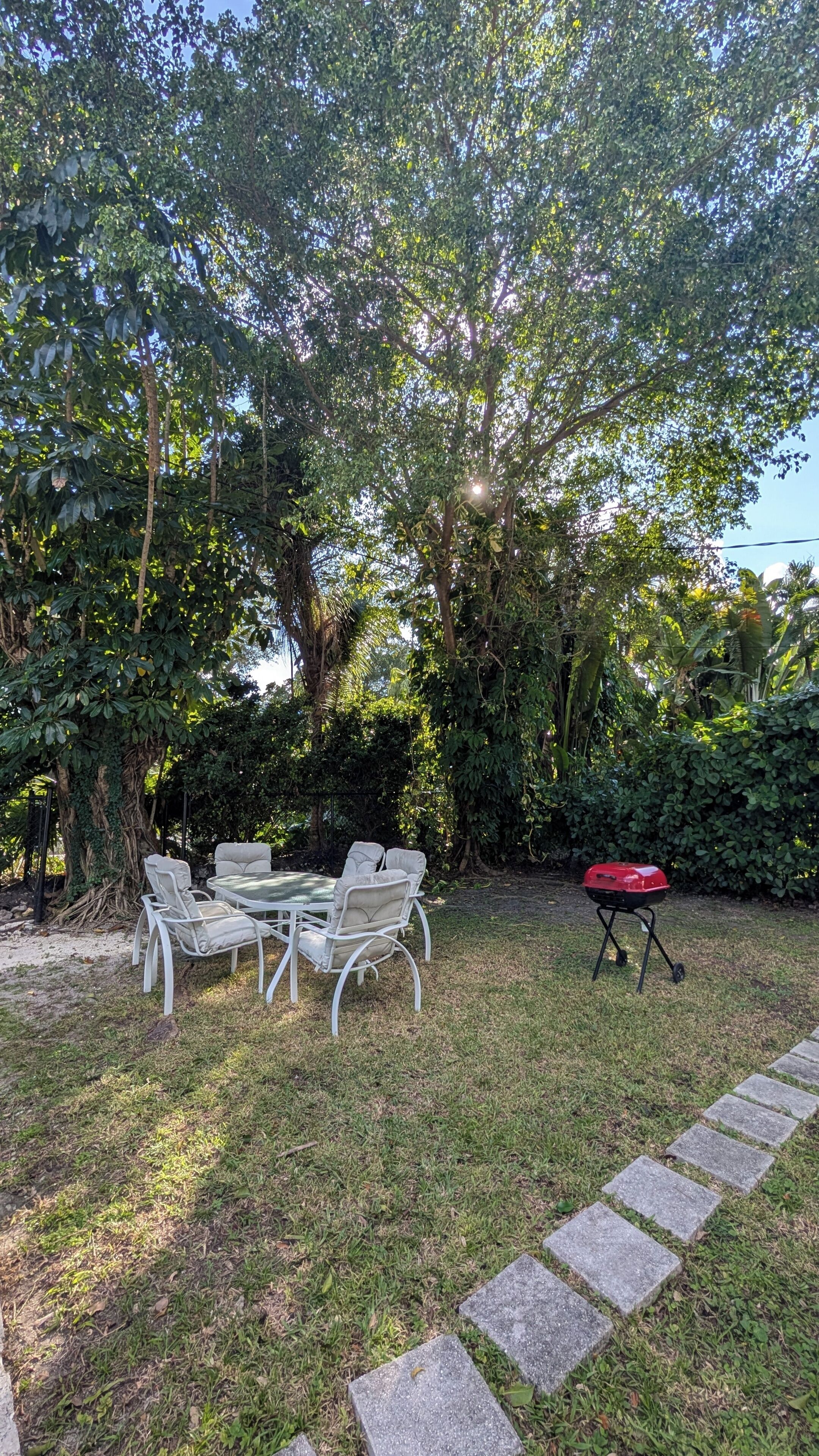Ein grünes Aussenareal mit einem Metalltisch und Stühlen, einem Grill und einem gepflasterten Weg.