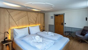 A hotel room with a large bed, a wooden headboard, a bedside table with a lamp, a chair, and a door. - Münsterhus Josefs Alpine Lodge - Three Towers Triple Room (Schruns)