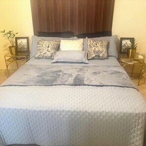A bed with a textured white cover, two bedside tables with framed pictures, and a wooden headboard.