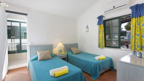 Une chambre avec deux lits, une table de chevet, une lampe, une fenêtre avec des rideaux, un appareil de climatisation et un petit élément de décoration murale.