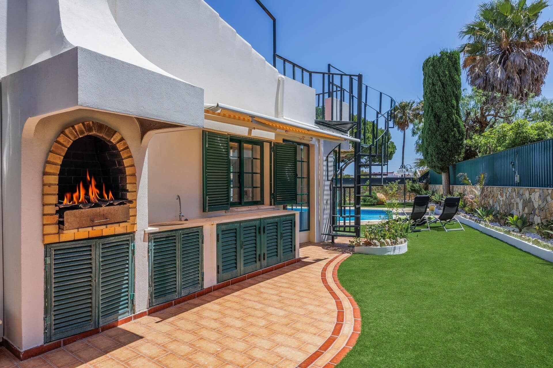 A modern outdoor kitchen with a brick fireplace, green shutters, and a pool area with lounge chairs.