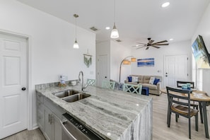 A kitchen with a marble countertop, stainless steel sink, and a dishwasher. There is a dining area with a table and chairs, a ceiling fan, and a painting on the wall.