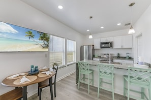 A modern kitchen with white cabinets, a stainless steel refrigerator, and a microwave. There is a dining area with a wooden table and chairs, and a large window with blinds.