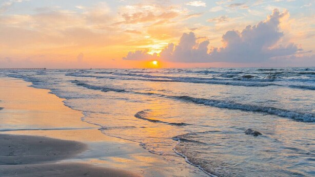 Ein Strand bei Sonnenuntergang mit Wellen, ein Pier in der Ferne und einem klaren Himmel.