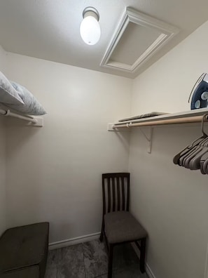 A small, enclosed laundry room with a chair, a shelf with hanging clothes, and an ironing board. - 1-bedroom studio with AC in vibrant TX (Austin)