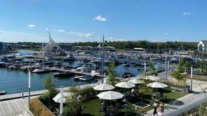 Un port de plaisance avec des bateaux amarrés, des tables et chaises à l’extérieur, protégées par des parasols, et un bâtiment moderne surmonté d’une flèche.