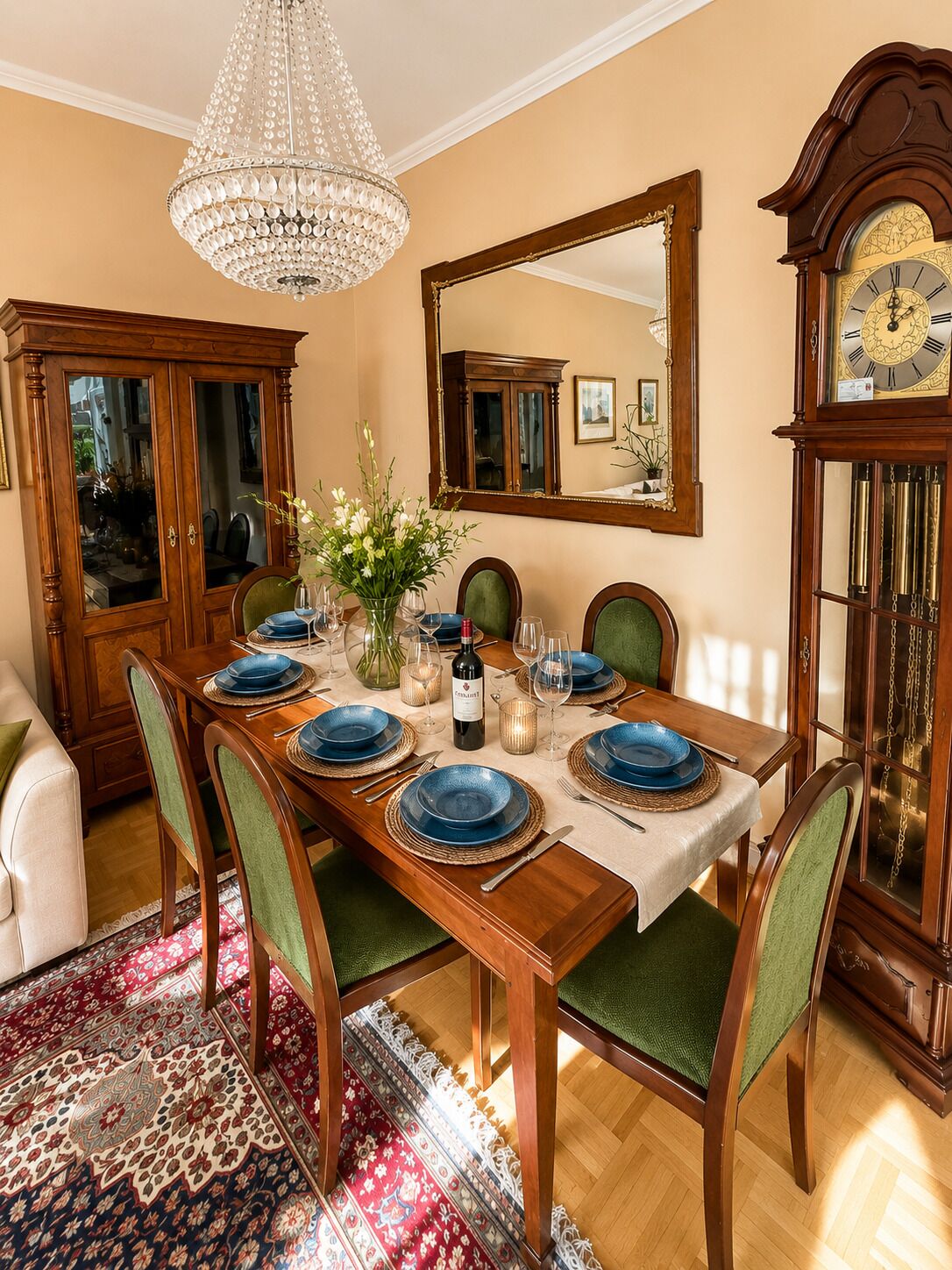 Ein Esszimmer mit einem Holztisch, gedeckt für sechs Personen, umgeben von Stühlen, einer Chandelüster, einem großen Spiegel und einer Standuhr.