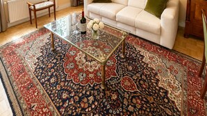 Una sala de estar con sofá, una mesa de centro de vidrio, un tapete estampado, una lámpara colgante y vistas de edificios a través de amplios ventanales.