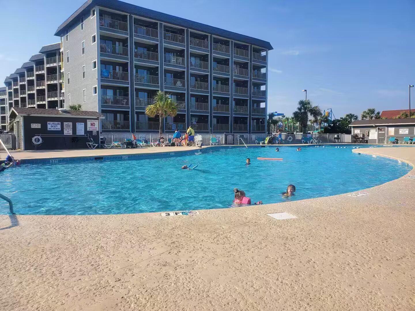 A swimming pool with multiple lanes, surrounded by a sandy beach and a multi-story building with balconies.