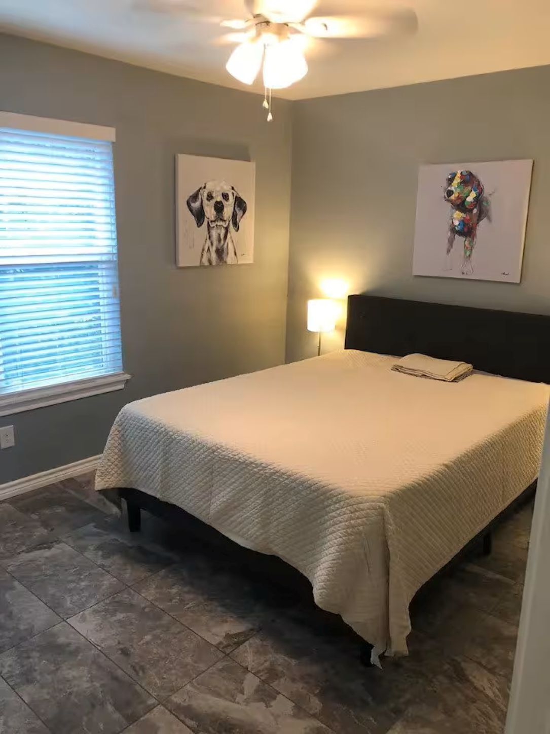 Une chambre à coucher comprenant un lit, un ventilateur de plafond, une fenêtre avec des stores et deux œuvres d’art murales.