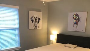 Un dormitorio con cama, ventilador de techo, ventana con persianas y dos cuadros en la pared.