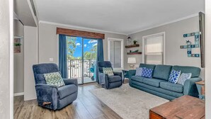 Ein Wohnzimmer mit einer Couch, zwei Sesseln, einem Couchtisch und einer Schiebeglastür, die auf einen Balkon führt.