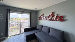 A room with a grey sofa, a large window with a view, and a wall with a red and white sign. - Perfect studio with AC in welcoming TX-3 (Austin)