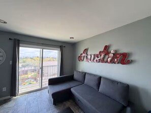 A room with a grey sofa, a large window with a view, and a wall with a red and white sign. - Perfect studio with AC in welcoming TX-3 (Austin)