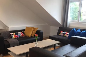 Ein modernes Wohnzimmer mit einer hohen Decke im Loft-Stil, zwei Sofas mit gemusterten Kissen, einem Couchtisch und einem Fenster mit Vorhängen.