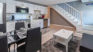 A modern kitchen with white cabinets, a dining area with a table and chairs, and a staircase leading to an upper level. - Spacious Home with Porch Sleeps 6 Near Pitt & UPMC (Pittsburgh)