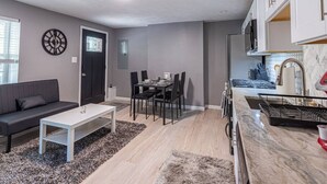 A modern living room with a sofa, dining table, and chairs, a kitchen with a stove and cabinets, and a clock on the wall. - Spacious Home with Porch Sleeps 6 Near Pitt & UPMC (Pittsburgh)
